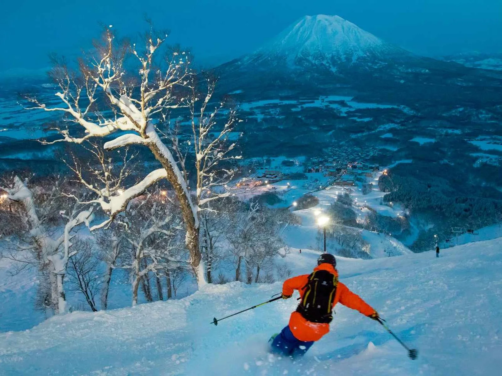 Esquiador de jaqueta laranja descendo uma pista nevada de Niseko com o monte Yotei ao fundo